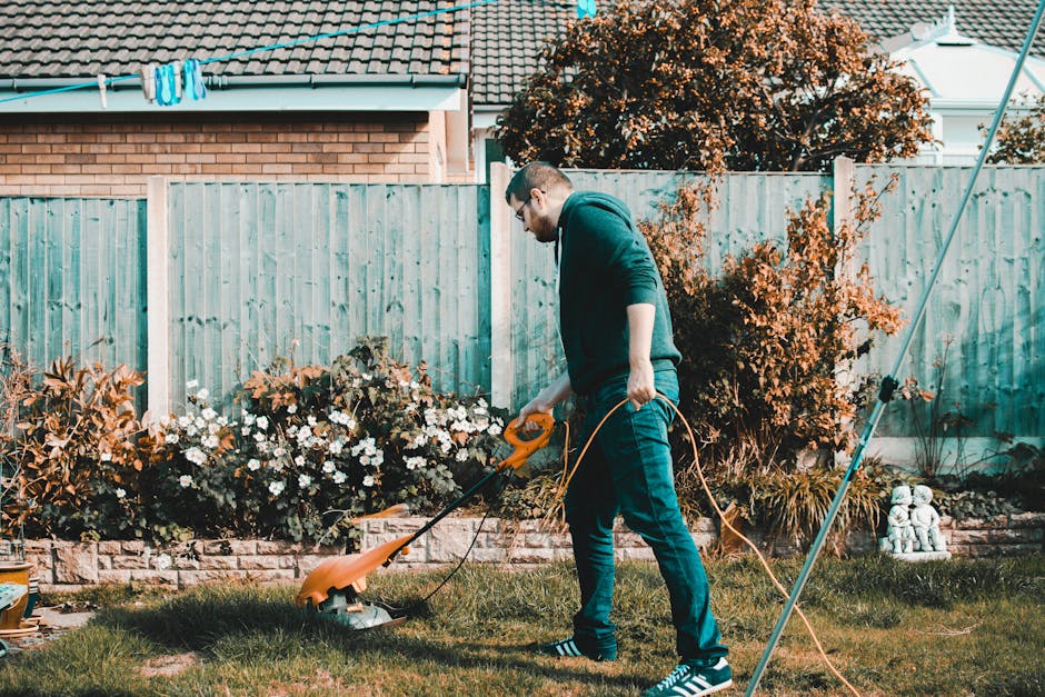 A man wearing glasses, a dark hoodie, and jeans is trimming the grass with a garden lawn mower in a backyard garden. The garden features a stone border, with flowering plants such as white daisies and